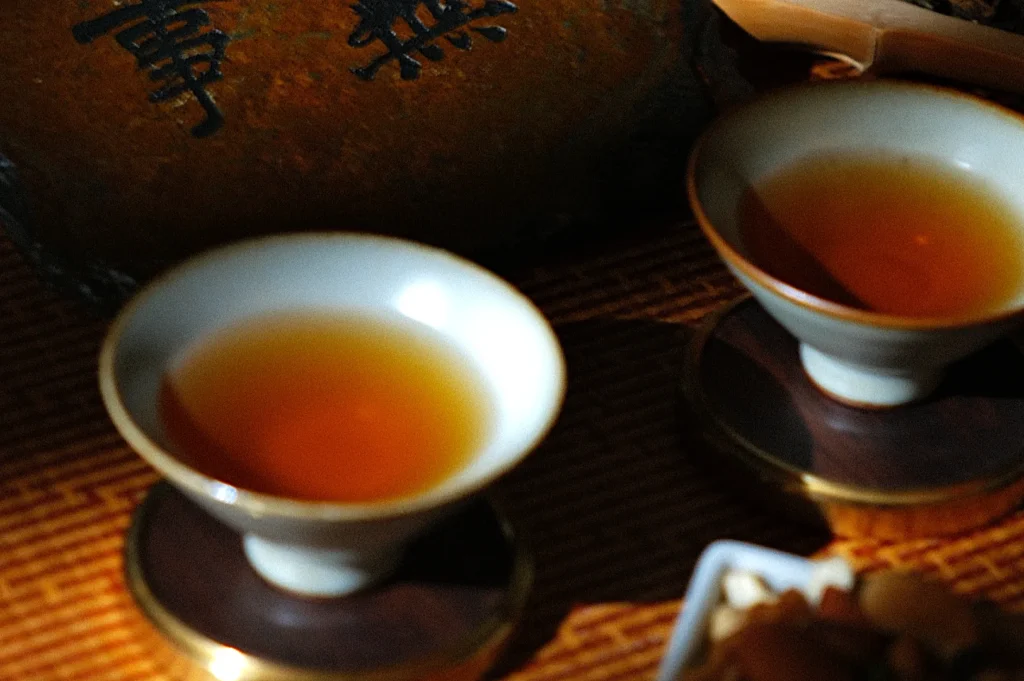 Two ceramic cups filled with amber-colored tea on a wooden saucer, set against a background with Chinese characters, creating a warm, serene atmosphere.