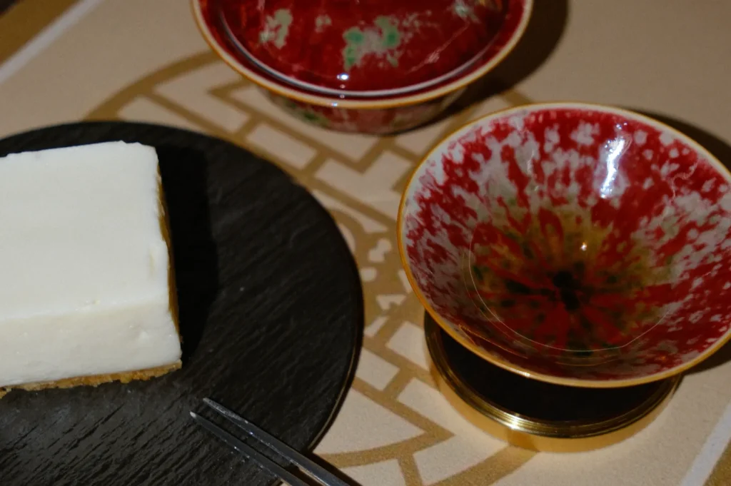 A piece of cheesecake sits on a dark slate plate with a two-pronged fork. Nearby, two ornate bowls feature a striking red and green pattern.