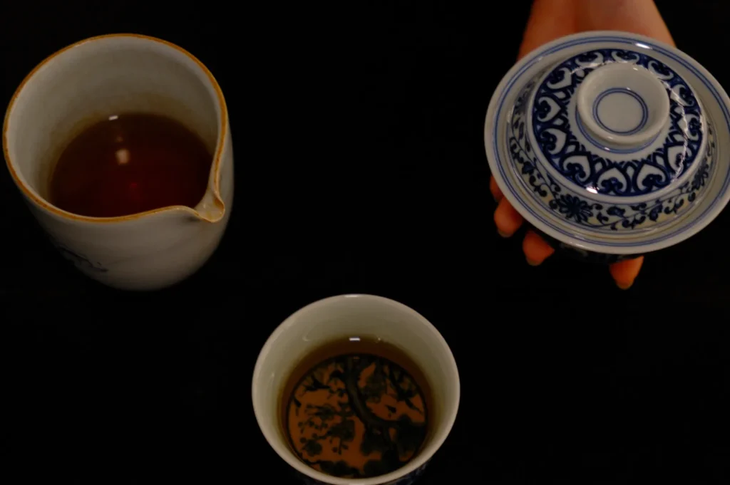 A hand holds a blue and white patterned lid above a traditional teacup, beside a small pitcher of tea on a black surface, creating a serene tea setting.