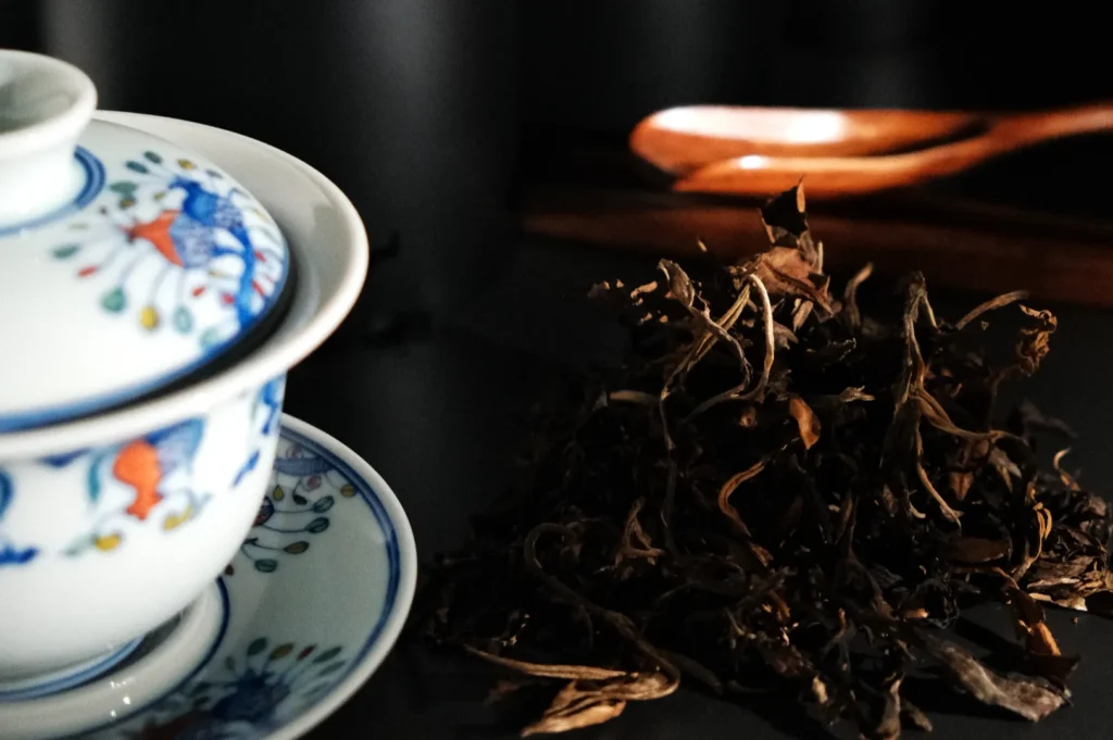 A cup and saucer with a decorative pattern beside a pile of loose black tea leaves and wooden utensils in the background.