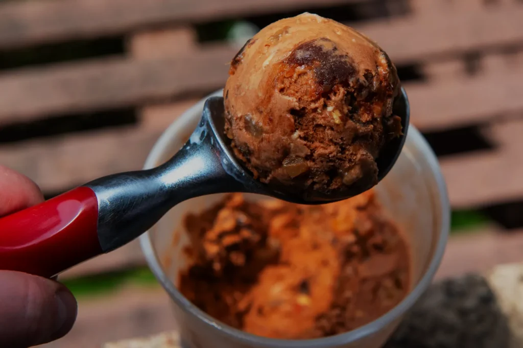 A hand holding an ice cream scoop with a scoop of chocolate ice cream above a partially empty container.