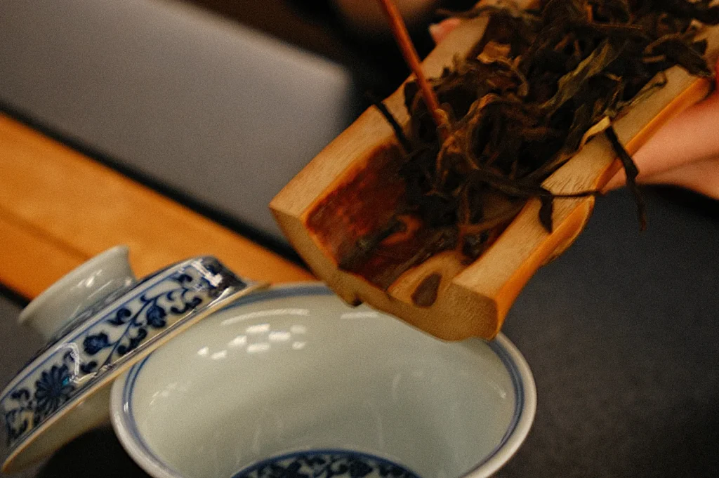 A person pouring loose tea leaves from a wooden tea caddy into a blue and white tea pot, with a traditional tea cup in the foreground.