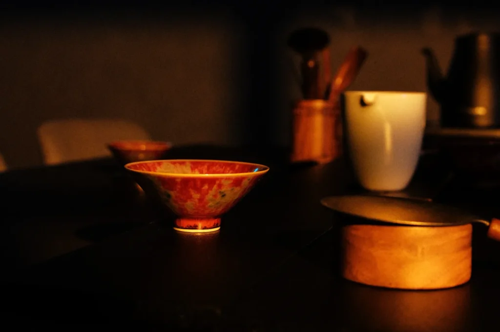A brightly colored bowl on a dark table with various kitchen utensils and a teapot in the background.