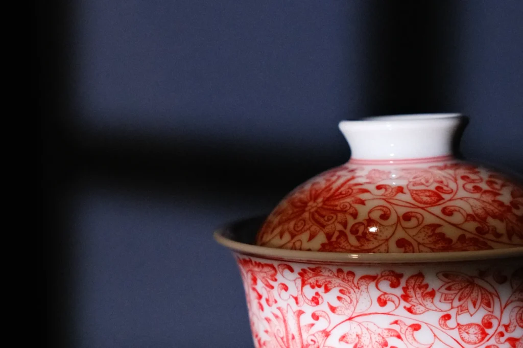 A close-up of a decorative bowl with a red floral pattern and a white lid against a dark background.