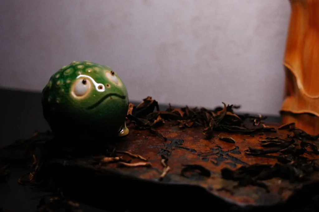A small green frog figurine sits on a surface covered with dried leaves.