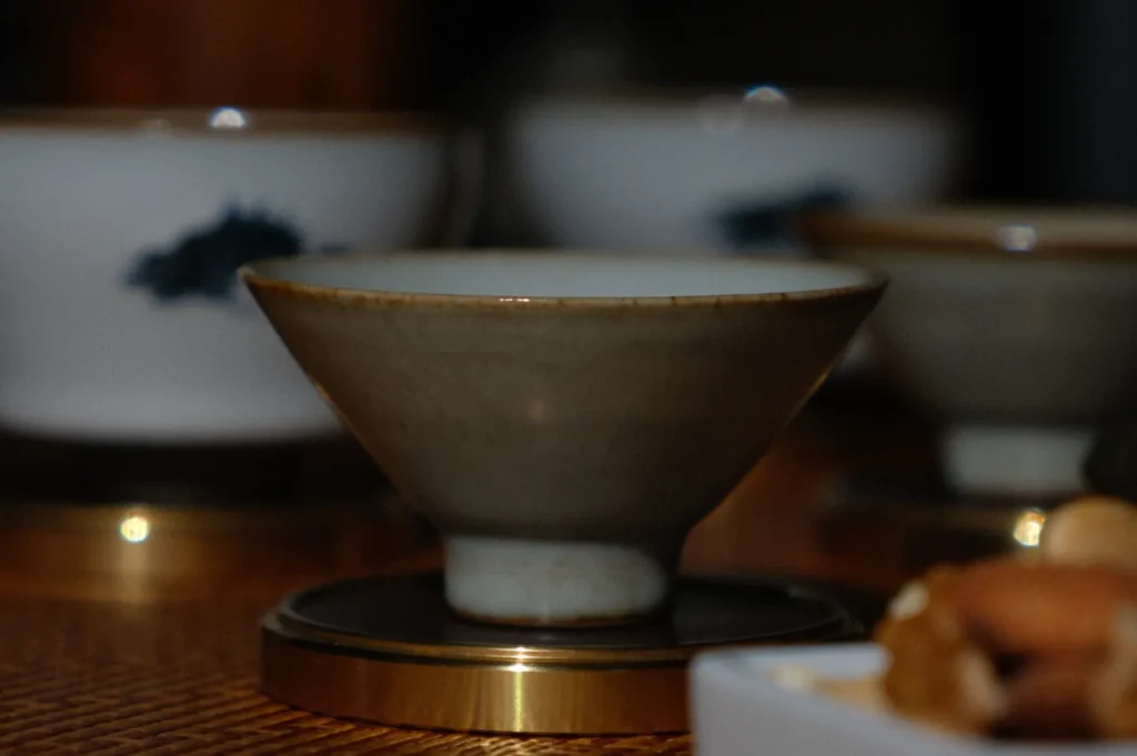 A close-up of a ceramic teacup on a wooden table, centered in focus. Surrounding cups are blurred, creating a cozy and intimate atmosphere.