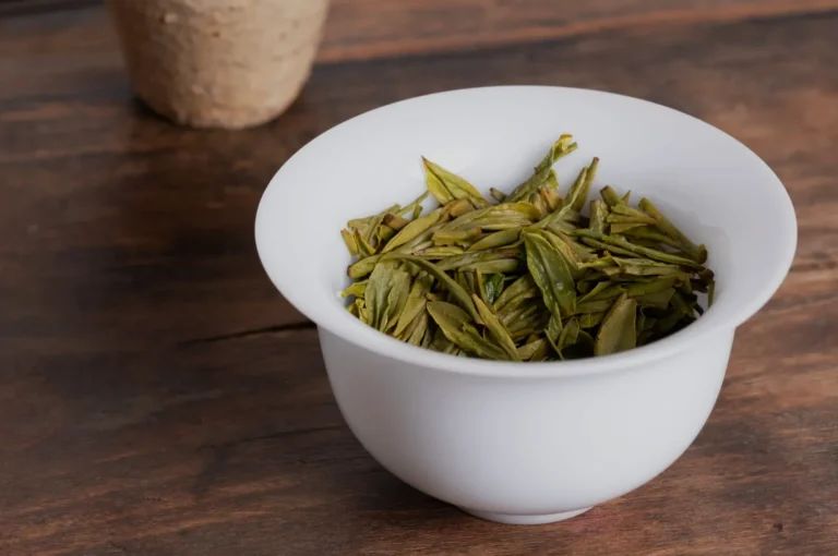 A white bowl filled with dried green tea leaves sitting on a wooden surface.
