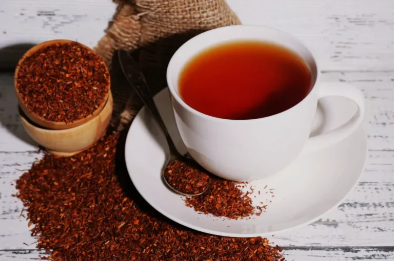 A cup of tea on a saucer with loose tea leaves spilled around it, along with a wooden bowl of tea leaves.