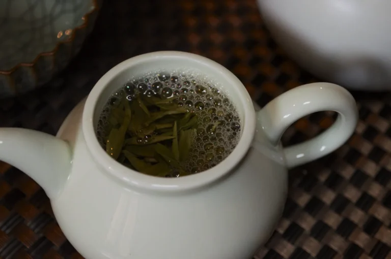 A small white teapot with freshly brewed green tea and tea leaves inside, placed on a woven mat.