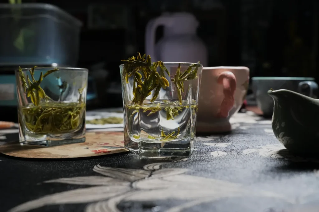 A close-up of a table with several glasses, some filled with green tea leaves submerged in water, and a variety of teaware in the background.