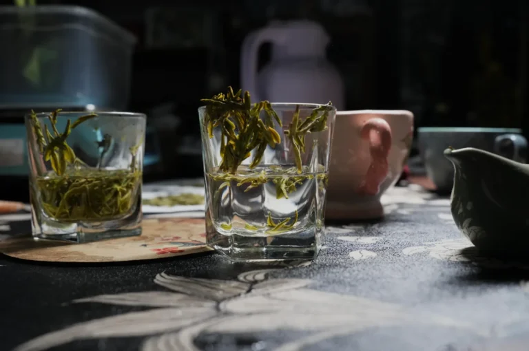 A close-up of a table with several glasses, some filled with green tea leaves submerged in water, and a variety of teaware in the background.