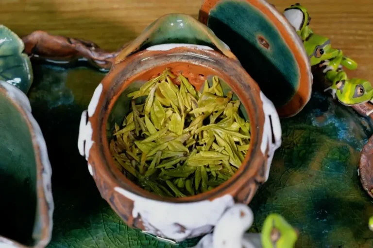A small clay pot with an open lid, containing green tea leaves, placed on a decorative surface.
