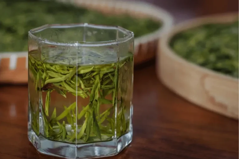 A glass of green tea with fresh tea leaves inside, placed on a wooden table, with dried tea leaves in the background.