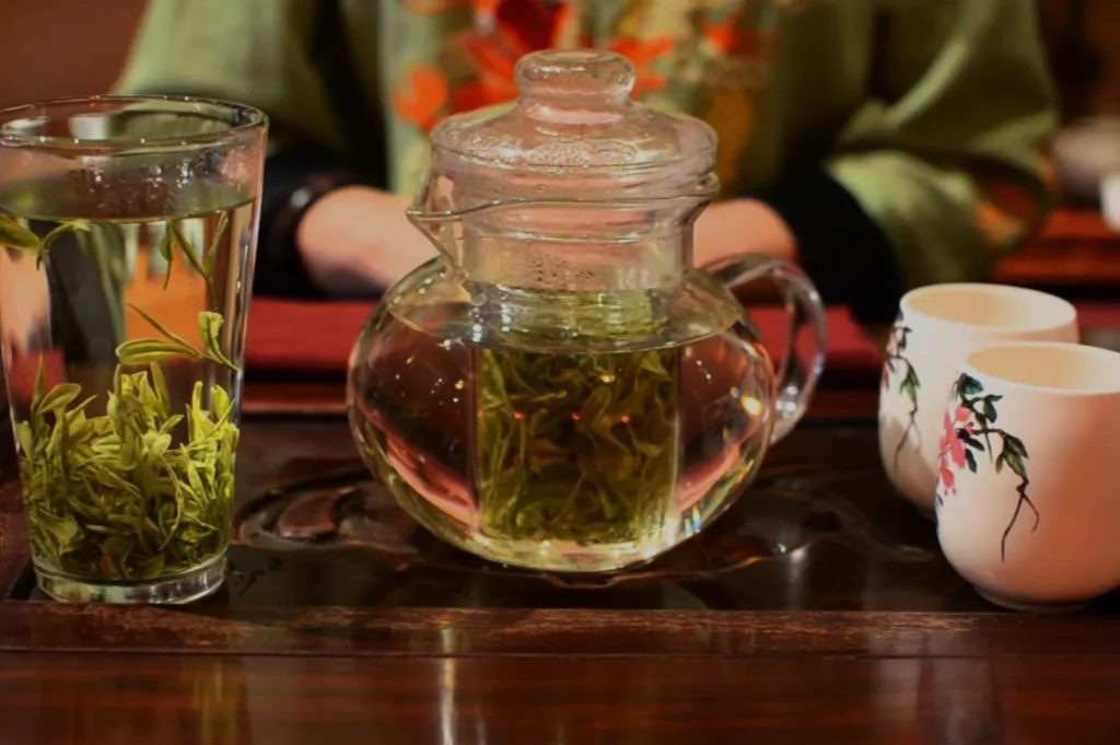 A glass teapot with green tea leaves, alongside two white tea cups and a glass filled with tea.