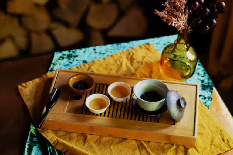 A wooden tray with tea cups, a tea pot, and a tea scoop, set on a colorful cloth with a vase of grapes in the background.