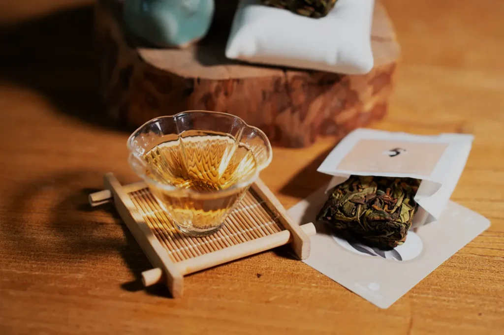 A small glass teacup with light amber tea, placed on a bamboo tray, next to a sachet of loose tea leaves and decorative items on a wooden surface.