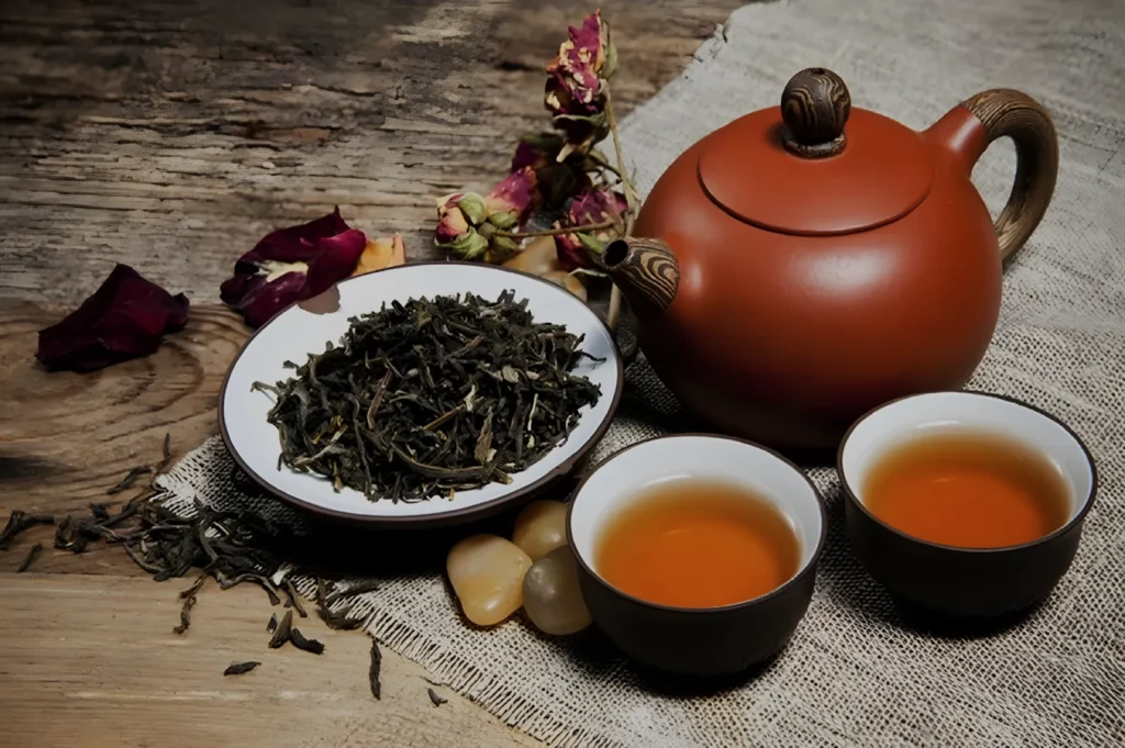 A teapot, a bowl of loose tea leaves, and two cups of brewed tea on a wooden surface, surrounded by dried rose petals and stone decorations.