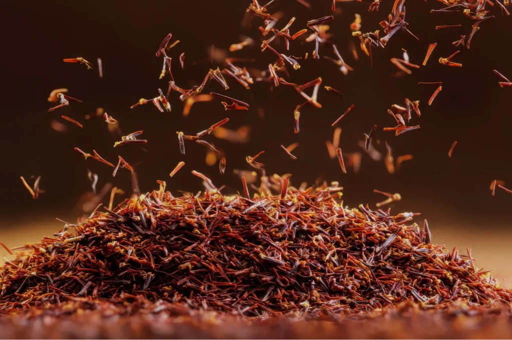 A close-up of a mound of saffron threads, with some threads appearing to float upwards against a blurred dark background.