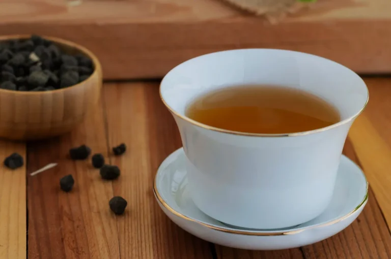 A white tea cup with a gold rim containing tea, placed on a saucer, with a wooden bowl of tea leaves in the background, surrounded by scattered tea leaves on a wooden surface.