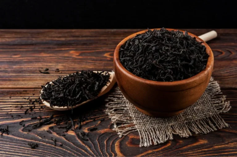 A wooden bowl filled with loose black tea leaves, accompanied by a wooden spoon holding some tea leaves, set on a textured burlap cloth on a rustic wooden surface.