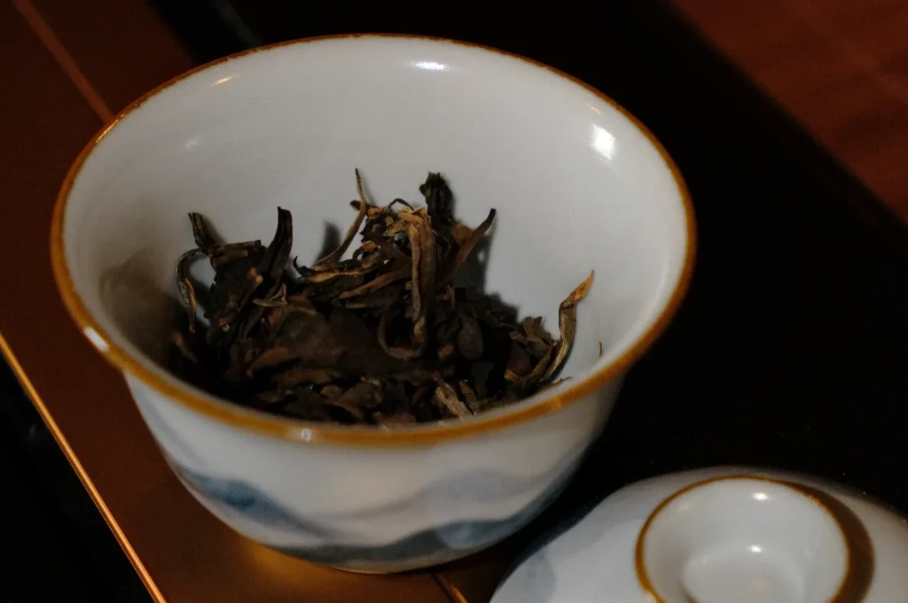 A small, ornate bowl containing loose tea leaves, with a lid beside it.
