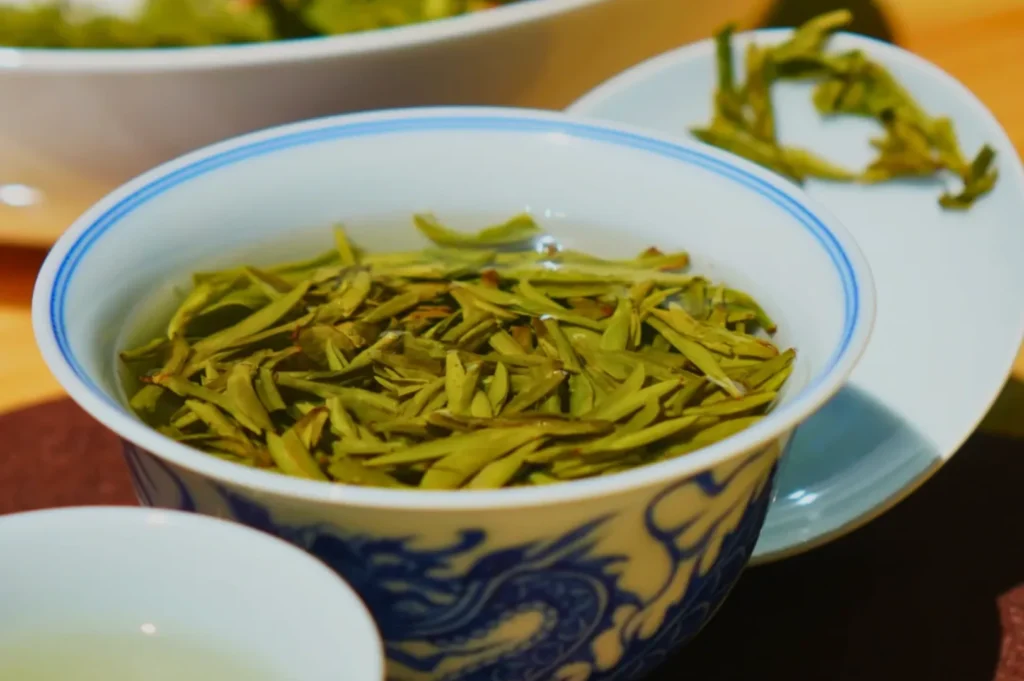 A bowl of loose green tea leaves with a small cup of tea beside it.