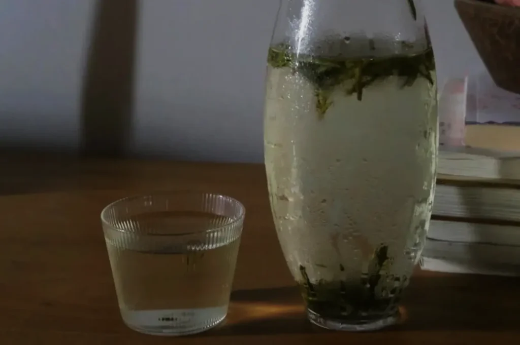 A glass of clear liquid with herbs beside a pitcher of the same liquid, placed on a wooden surface with books in the background.