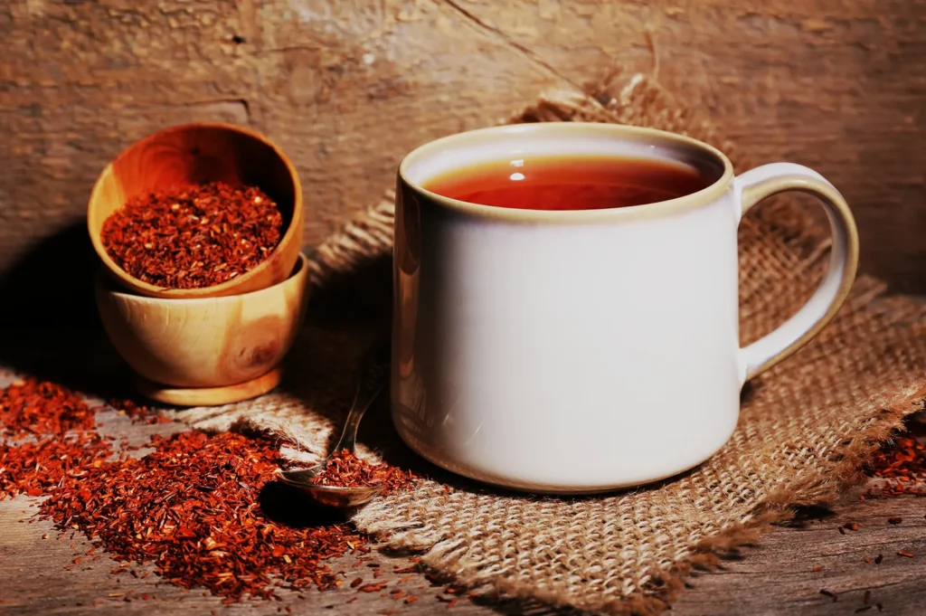 A white mug filled with tea next to a wooden bowl of spices and some scattered spices on a burlap surface.