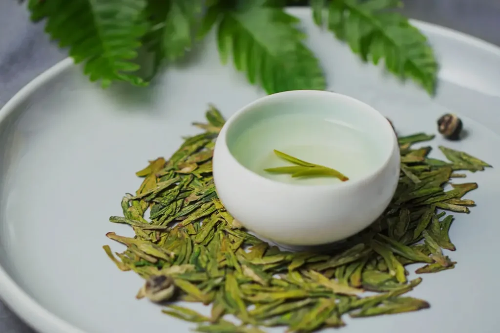 A small white bowl containing a clear liquid is placed on a plate with dried green tea leaves and decorative elements like a small green leaf and a few stones.