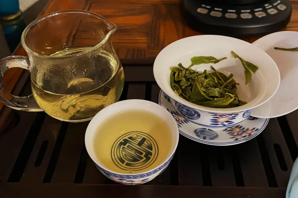 A glass teapot with steeped tea next to a bowl of tea leaves and a small cup of tea.