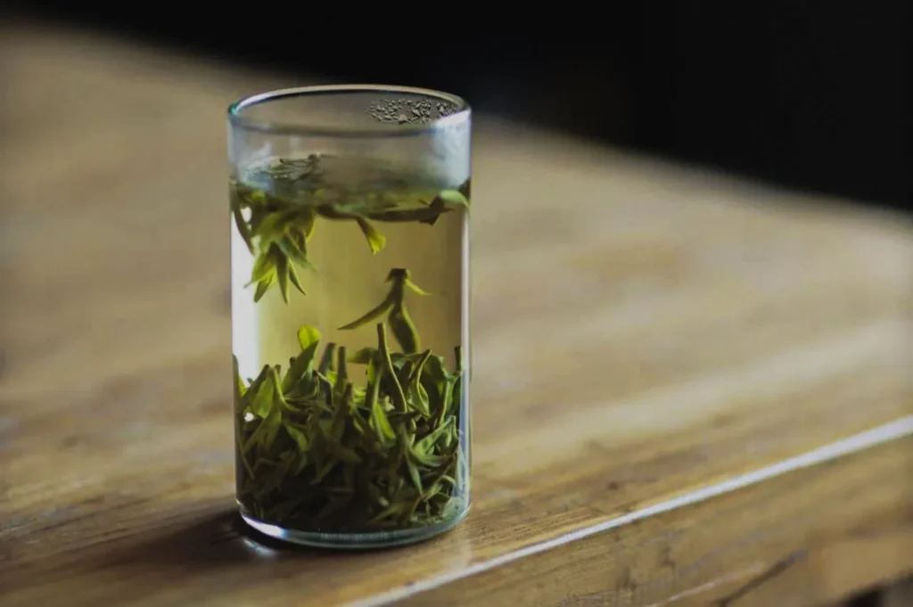 A glass of tea with green tea leaves steeping in it, sitting on a wooden table.