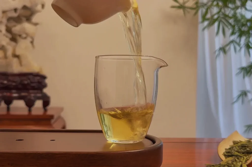 A teapot pouring green tea into a glass cup on a wooden tray with a blurred background of bamboo.
