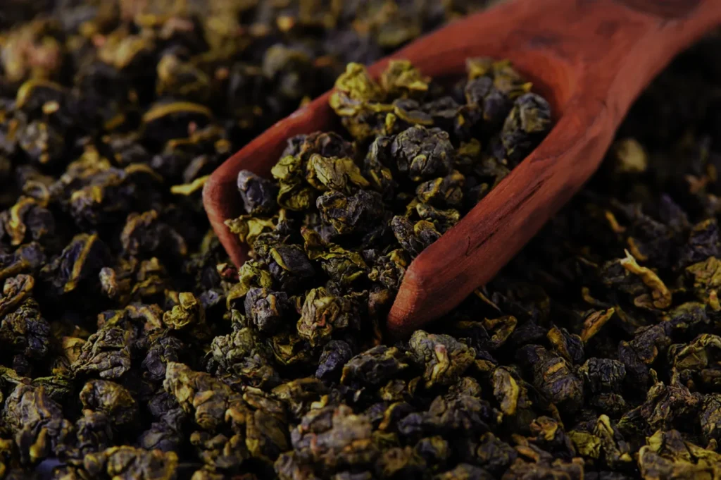 A wooden scoop filled with dark green and purple oolong tea leaves, resting on a bed of loose tea leaves.