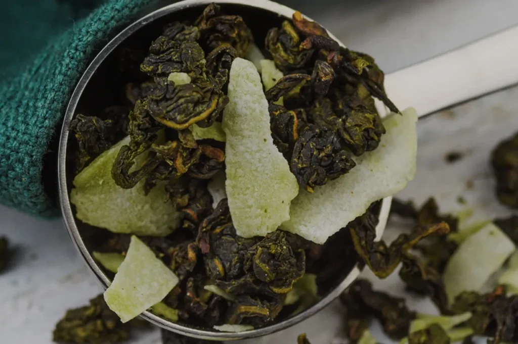 A metal measuring spoon filled with dried green tea leaves and apple pieces.