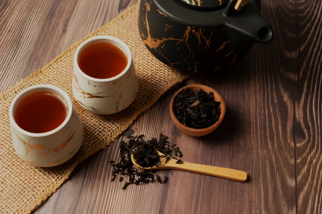 A teapot with two cups of tea, a small bowl of loose tea leaves, and a wooden spoon on a textured surface.