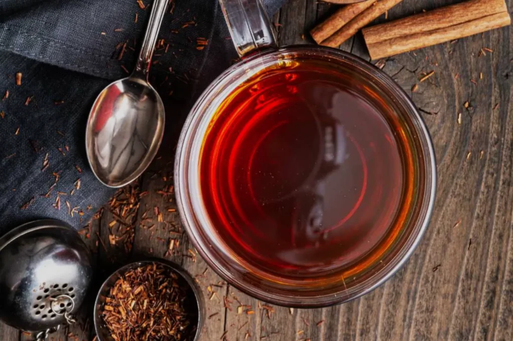 A cup of dark tea placed on a wooden surface, with loose tea leaves and cinnamon sticks nearby.