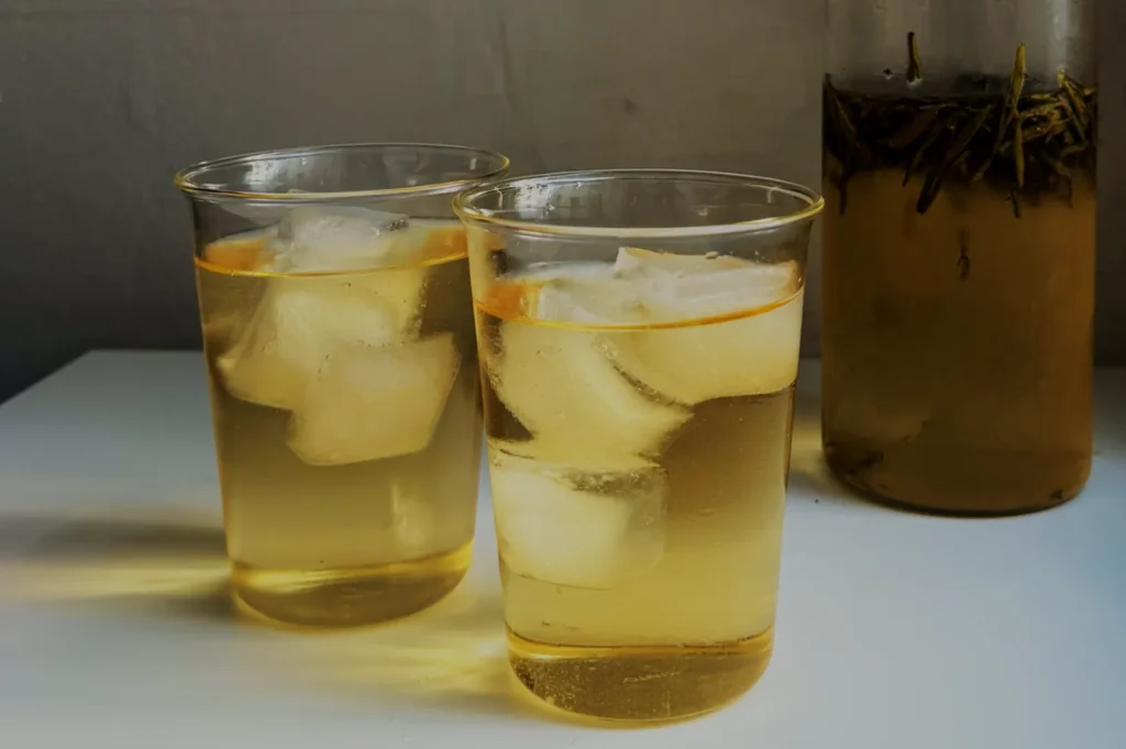 Two glasses filled with a light-colored drink and ice, placed on a white surface, next to a glass jar containing a darker beverage with visible ingredients.
