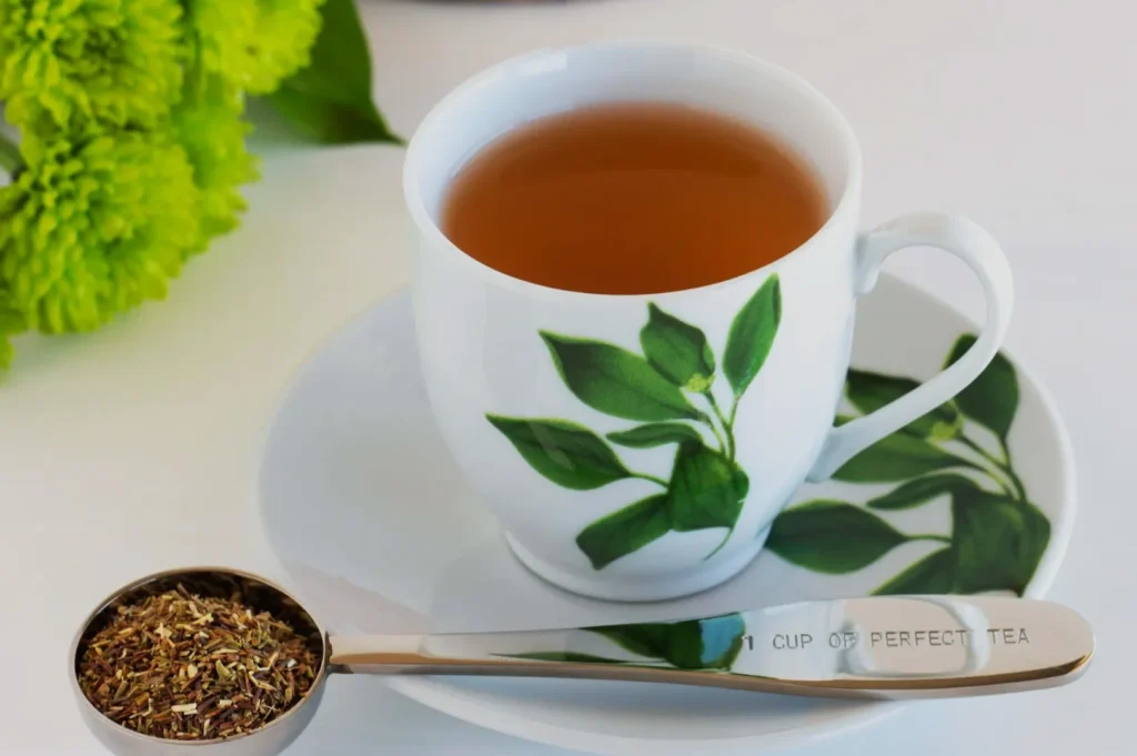 A cup of tea with a spoon of loose tea leaves beside it, accompanied by green flowers in the background.