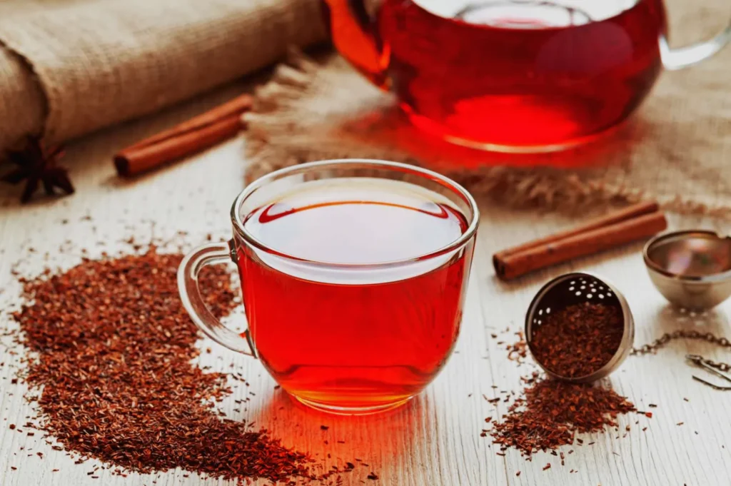A cup of red tea next to a teapot, with loose tea leaves and a tea infuser on a wooden surface.