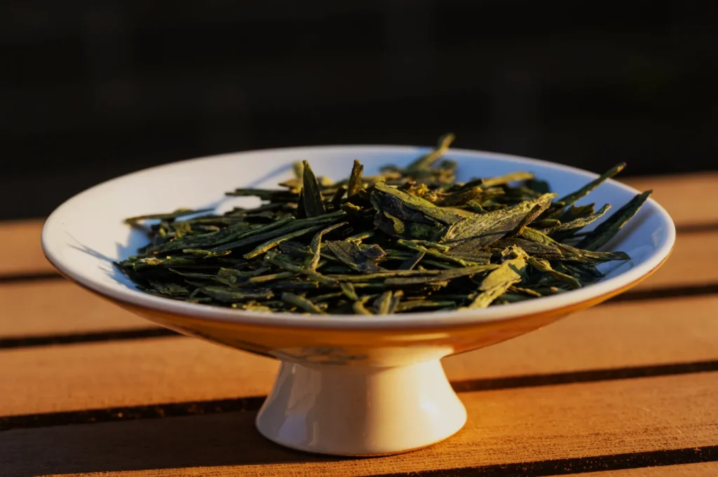 A small white bowl filled with green loose tea leaves, sitting on a wooden surface.