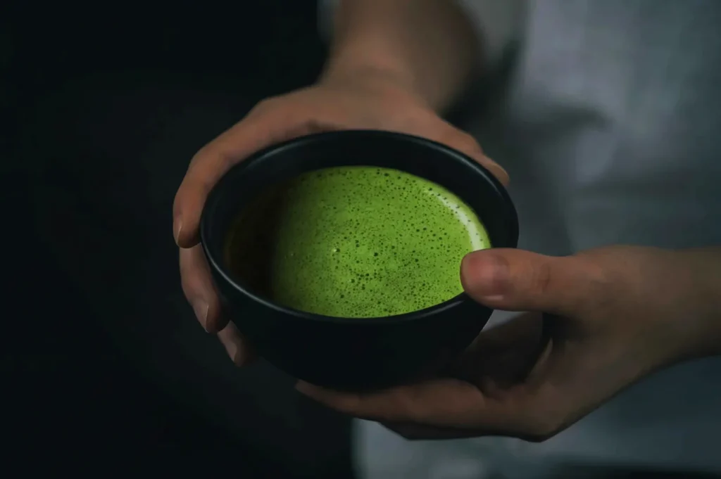 A person holding a black bowl containing green frothy liquid, possibly matcha.