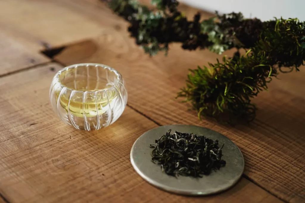 A small glass cup of tea next to a mound of loose tea leaves and some mossy twigs on a wooden surface.