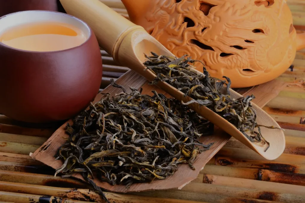 A wooden scoop of loose tea leaves next to a brown teacup and a tea pot on a bamboo mat.
