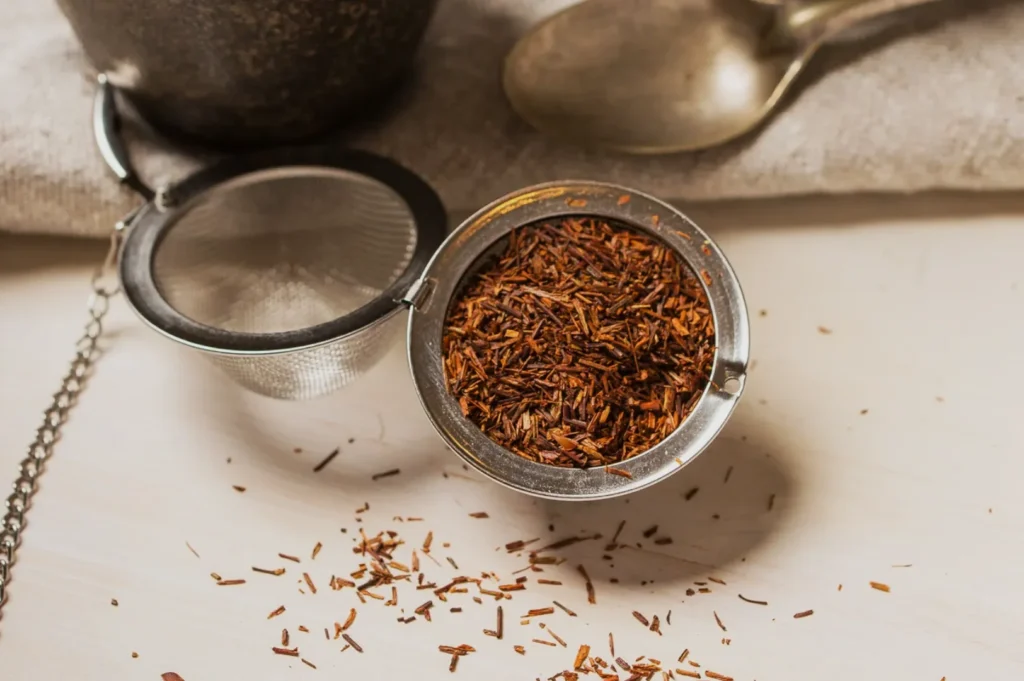 A close-up of a tea infuser filled with loose rooibos tea leaves, with some leaves scattered around it, and a spoon in the background.