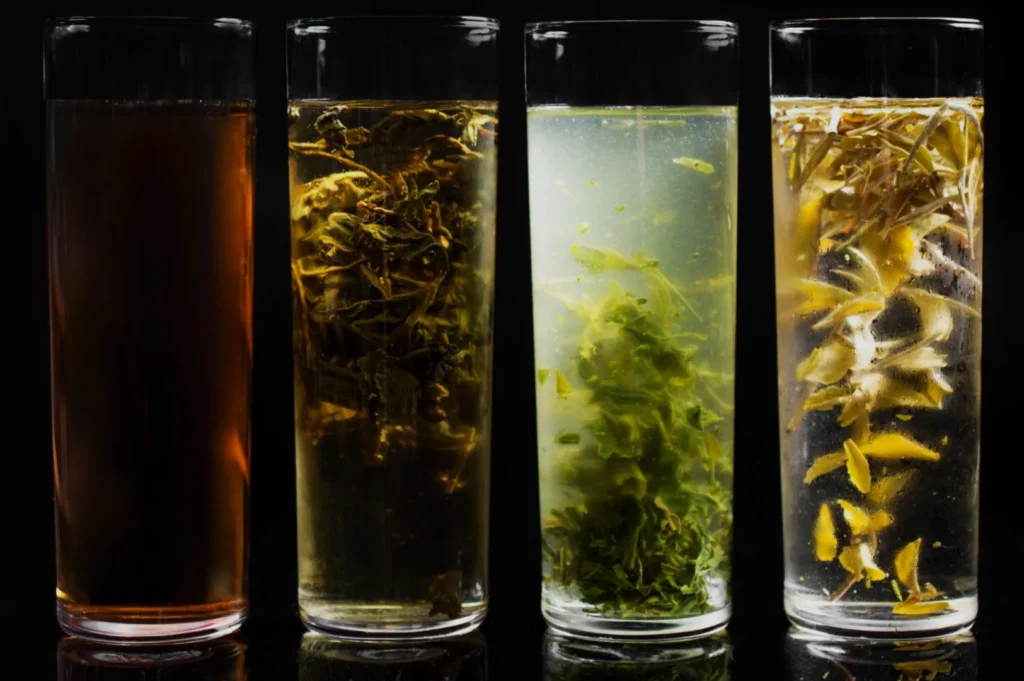 Four tall glasses of differently colored teas or herbal infusions, ranging from dark to light, with visible tea leaves and herbs inside.