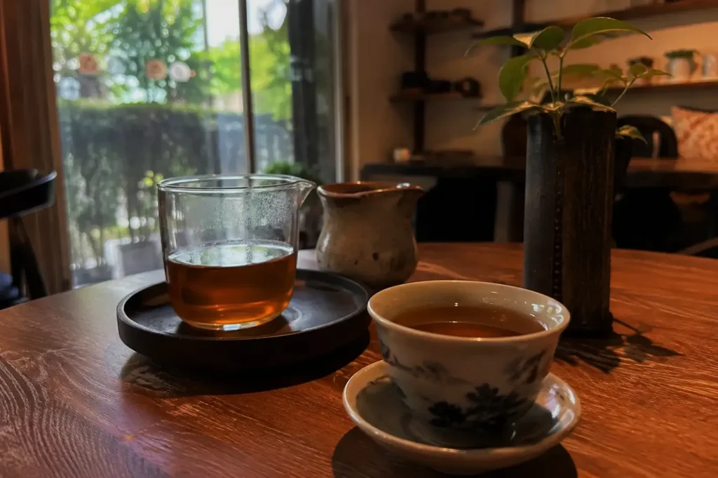 A cup of tea on a wooden table, alongside a glass of tea and a small pitcher, with a window showing greenery in the background.