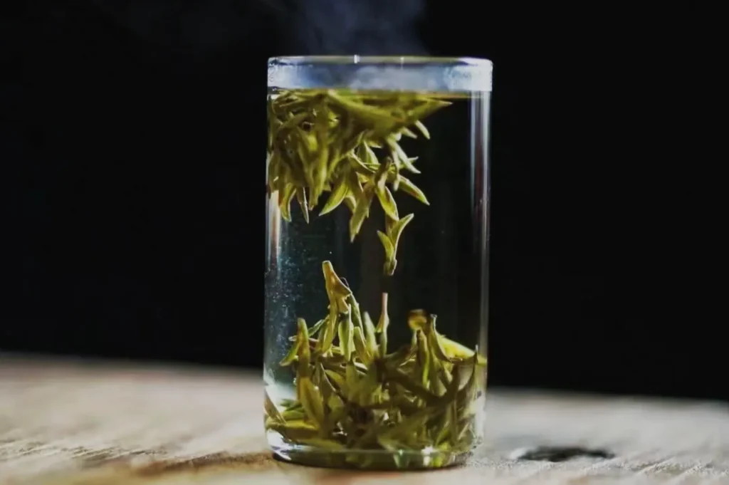 A clear glass filled with water and green tea leaves floating inside. Steam is rising from the glass against a dark background.