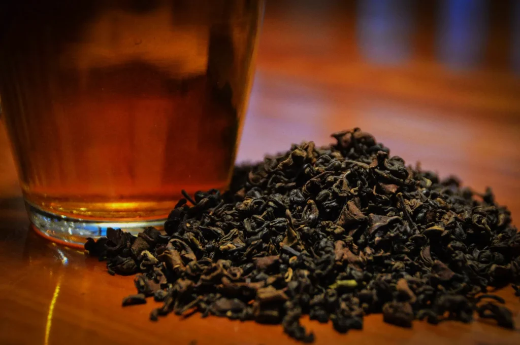 A glass of amber liquid next to a mound of loose black tea leaves on a wooden surface.