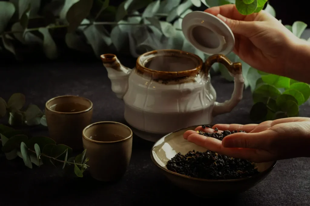 A person's hand pouring loose tea leaves from a bowl into a teapot, with two empty teacups and greenery in the background.