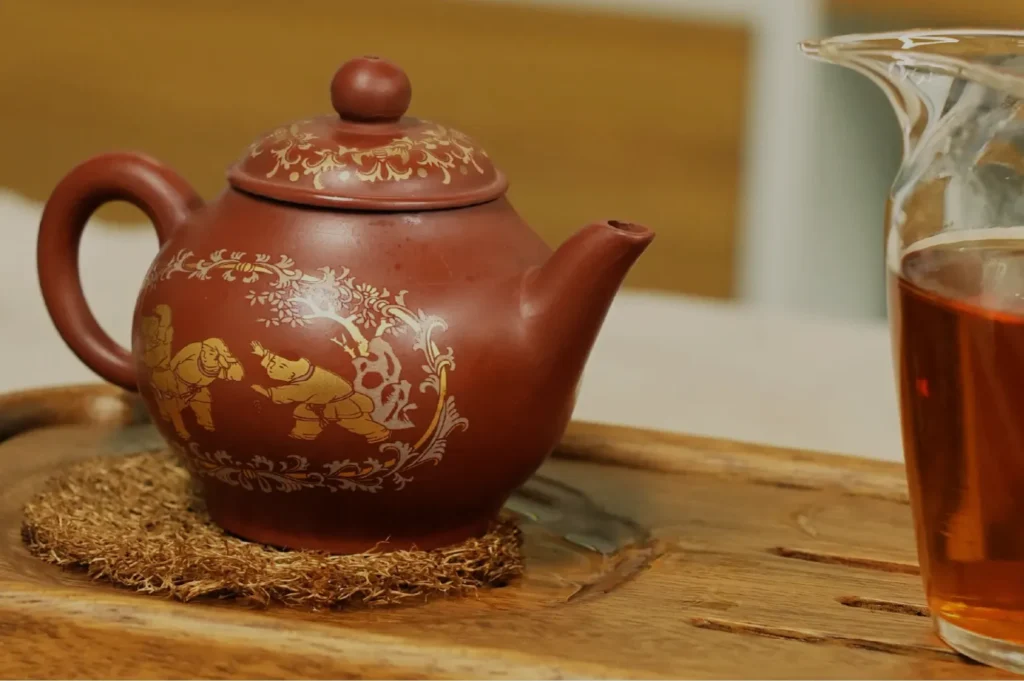 A brown ceramic teapot with decorative floral patterns, placed on a wooden tray alongside a glass of tea.